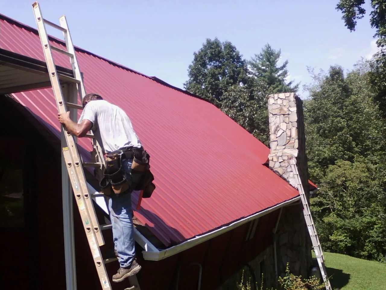 Roofer on a ladder beside a red metal roof and stone chimney — Soffit & Fascia Repair by Roofing Brothers in Solway